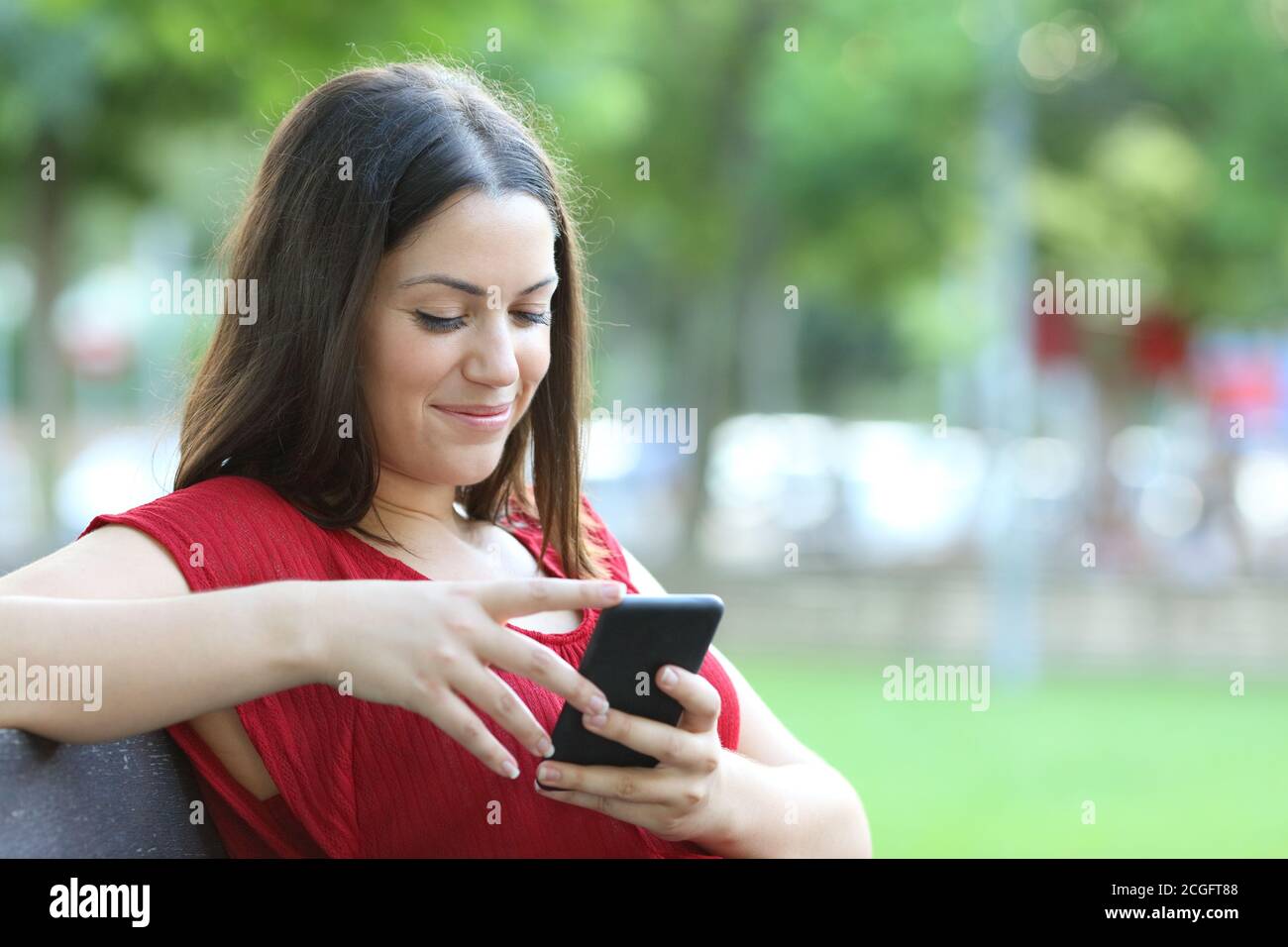 Donna sorridente in rosso controlla lo smartphone seduto su un panca in un parco Foto Stock