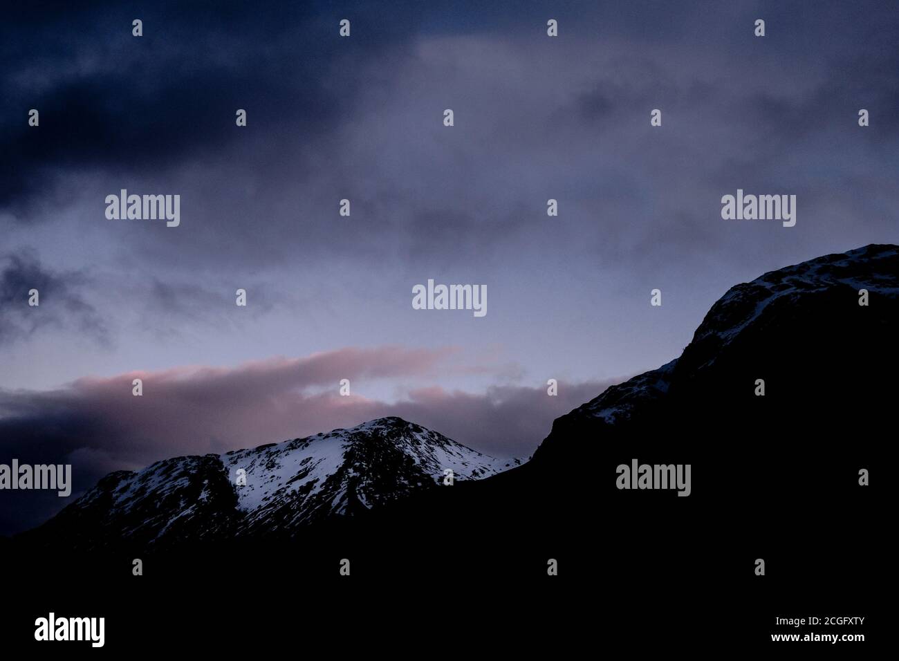 Vista delle montagne innevate di Glen Coe, Scozia, alle capanne del tramonto prima che scenda l'oscurità, con un po' di colore dal tramonto nel cielo. Foto Stock