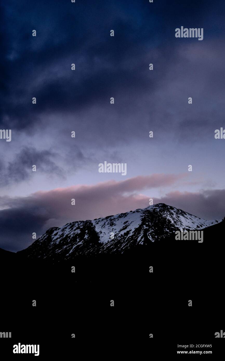 Vista delle montagne innevate di Glen Coe, Scozia, alle capanne del tramonto prima che scenda l'oscurità, con un po' di colore dal tramonto nel cielo. Foto Stock