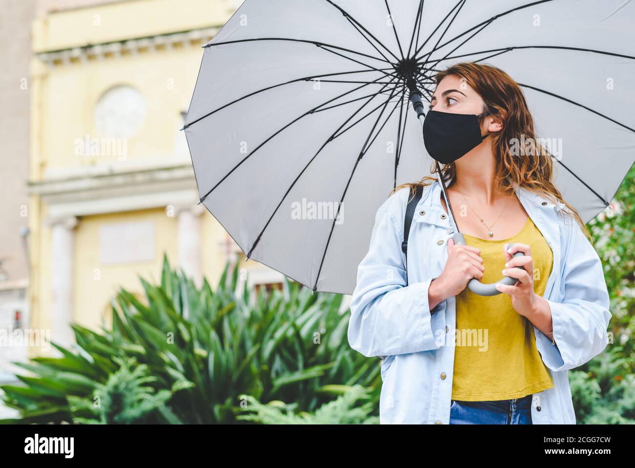 bella giovane donna dai capelli rossi che indossa un mash nero igienico in piedi in una strada e tenendo un ombrello con entrambi mani su una pioggia inizio autunno da Foto Stock