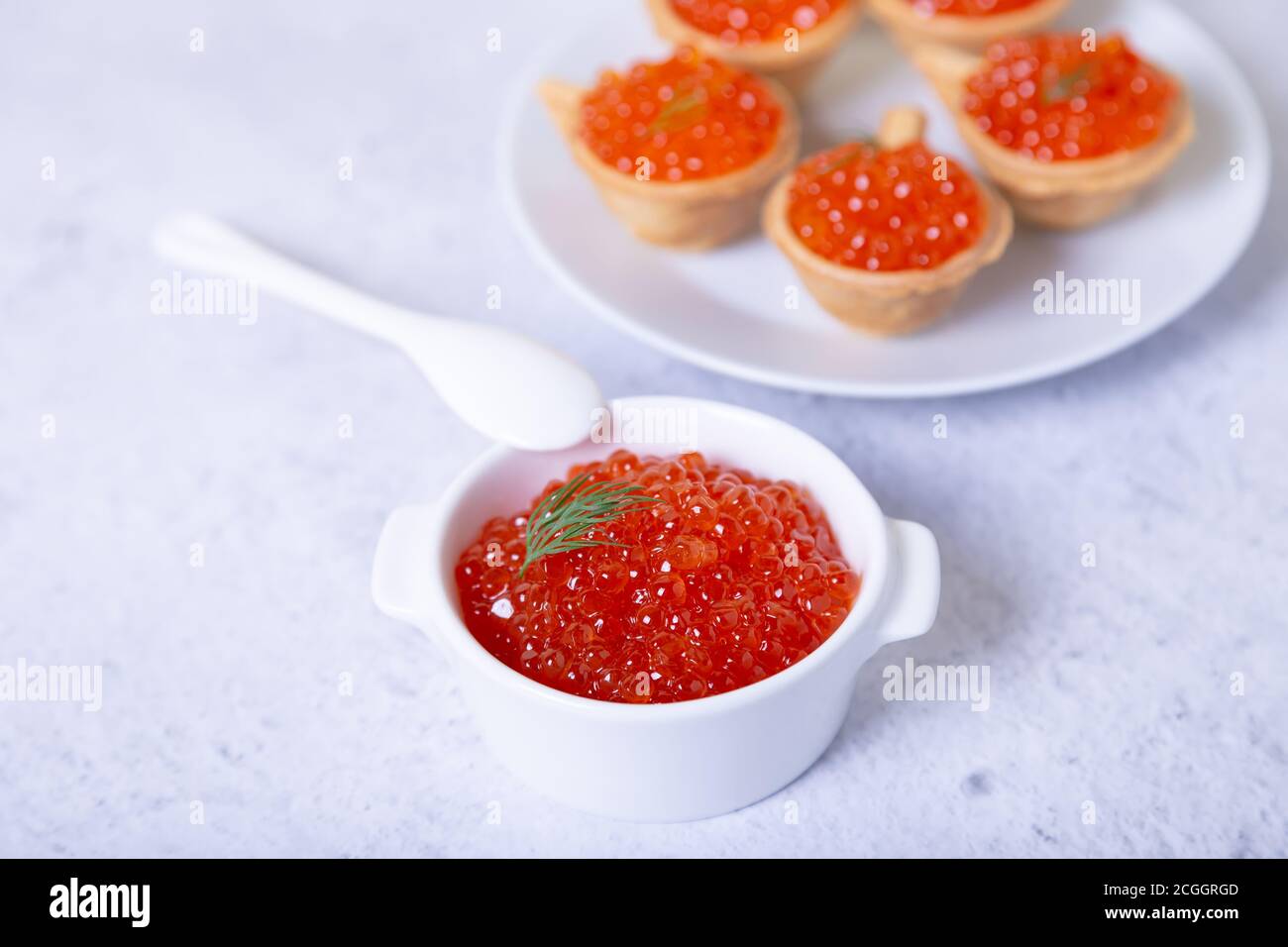 Caviale rosso (caviale di salmone) in una tazza bianca. Sullo sfondo è presente un piatto con tartine e caviale. Messa a fuoco selettiva, primo piano. Foto Stock