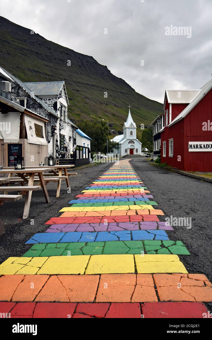 La strada arcobaleno dipinta a Seydisfjordur in un giorno di colore grigio. Eastfjords, Islanda. Tavoli da picnic sono all'esterno di un ristorante vicino alla strada. Foto Stock