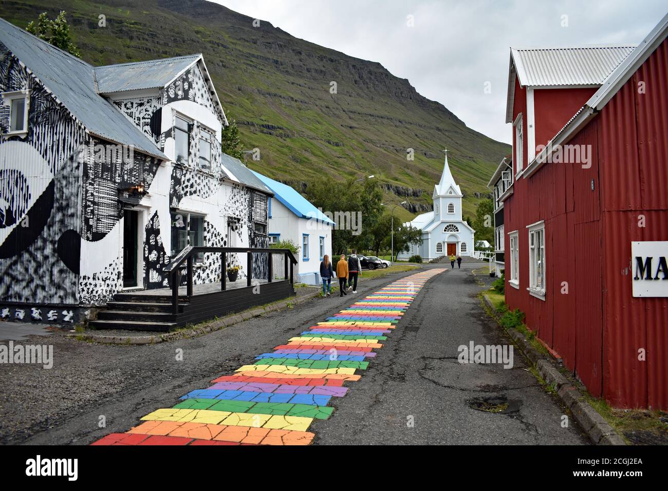 I visitatori camminano accanto a un negozio di motivi bianchi e neri lungo la strada arcobaleno dipinta a Seydisfjordur verso la chiesa. Eastfjords, Islanda Foto Stock