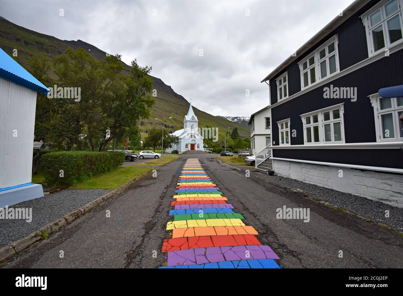 Guardando lungo la colorata strada arcobaleno dipinto a Seydisfjordur verso la chiesa e le montagne in un giorno coperto, grigio. Eastfjords, Islanda Foto Stock