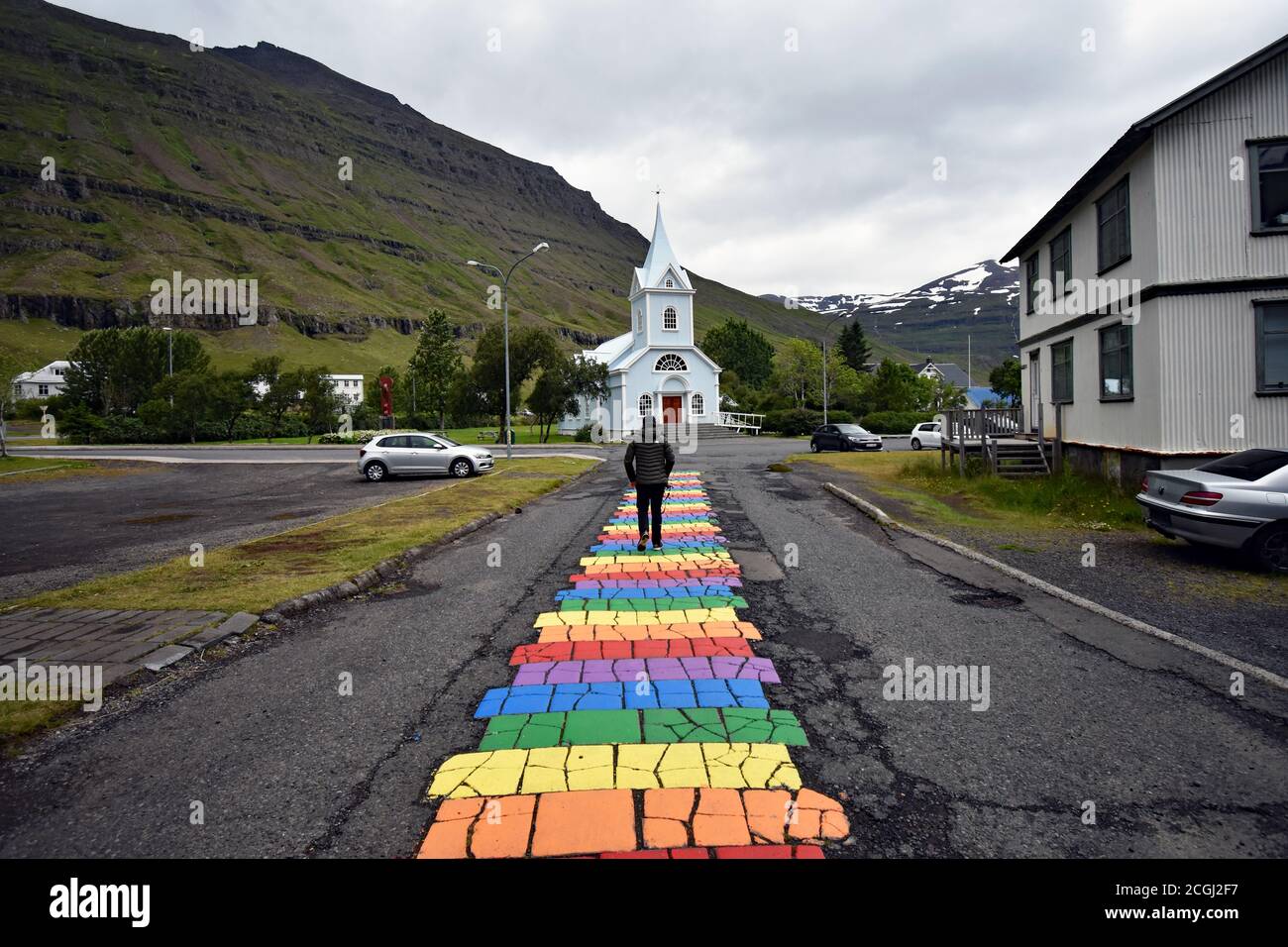 Un turista maschile cammina lungo la strada arcobaleno dipinta in Seydisfjordur verso la chiesa e le montagne in un giorno sovrastato, grigio. Eastfjords, Islanda Foto Stock