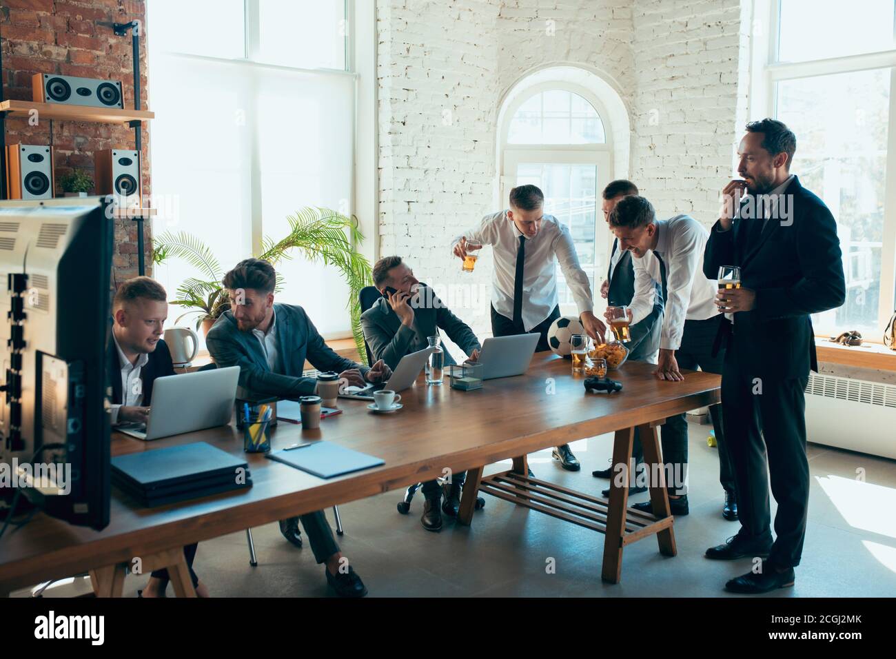 Guardare il calcio, bere birra, rallegrarsi. I colleghi sono felici e spensierati e si divertono in ufficio mentre i colleghi lavorano sodo e altamente concentrati. Concetto di divertimento, riposo, occupazione professionale. Foto Stock