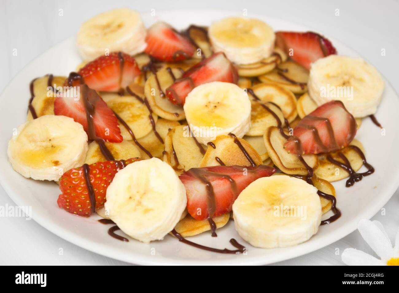 Cibo alla moda - cereali pancake. Un mucchio di mini frittelle di cereali su un piatto bianco su sfondo bianco. Fette di fragola su un piatto, condite con sciroppo. Foto Stock