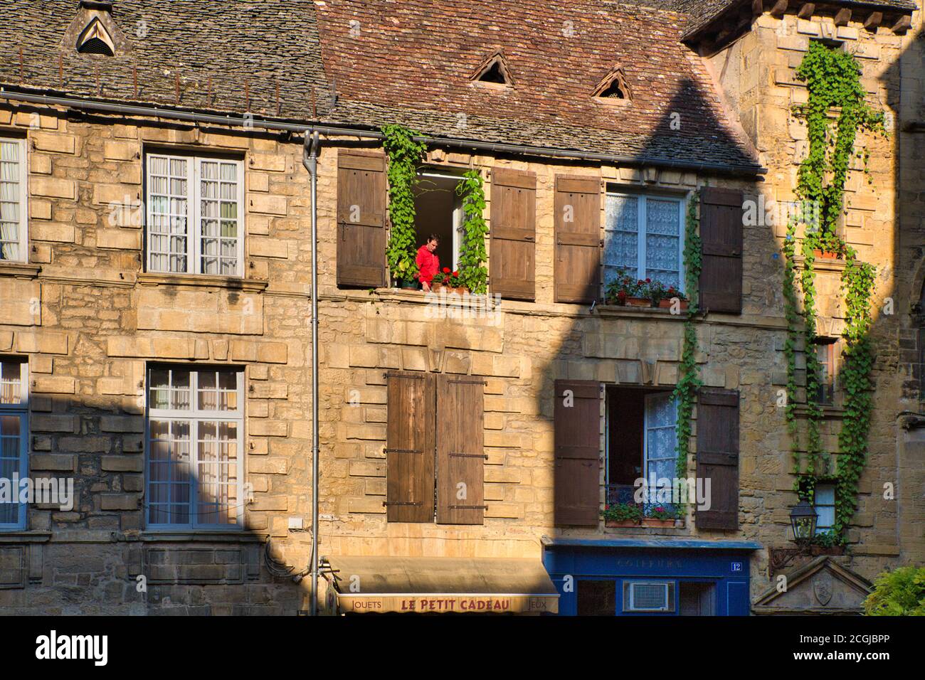 Una persona in maglietta rossa incorniciata nella finestra aperta della loro casa con fogliame e persiane visibili, Languedoc-Roussillon, Francia Foto Stock