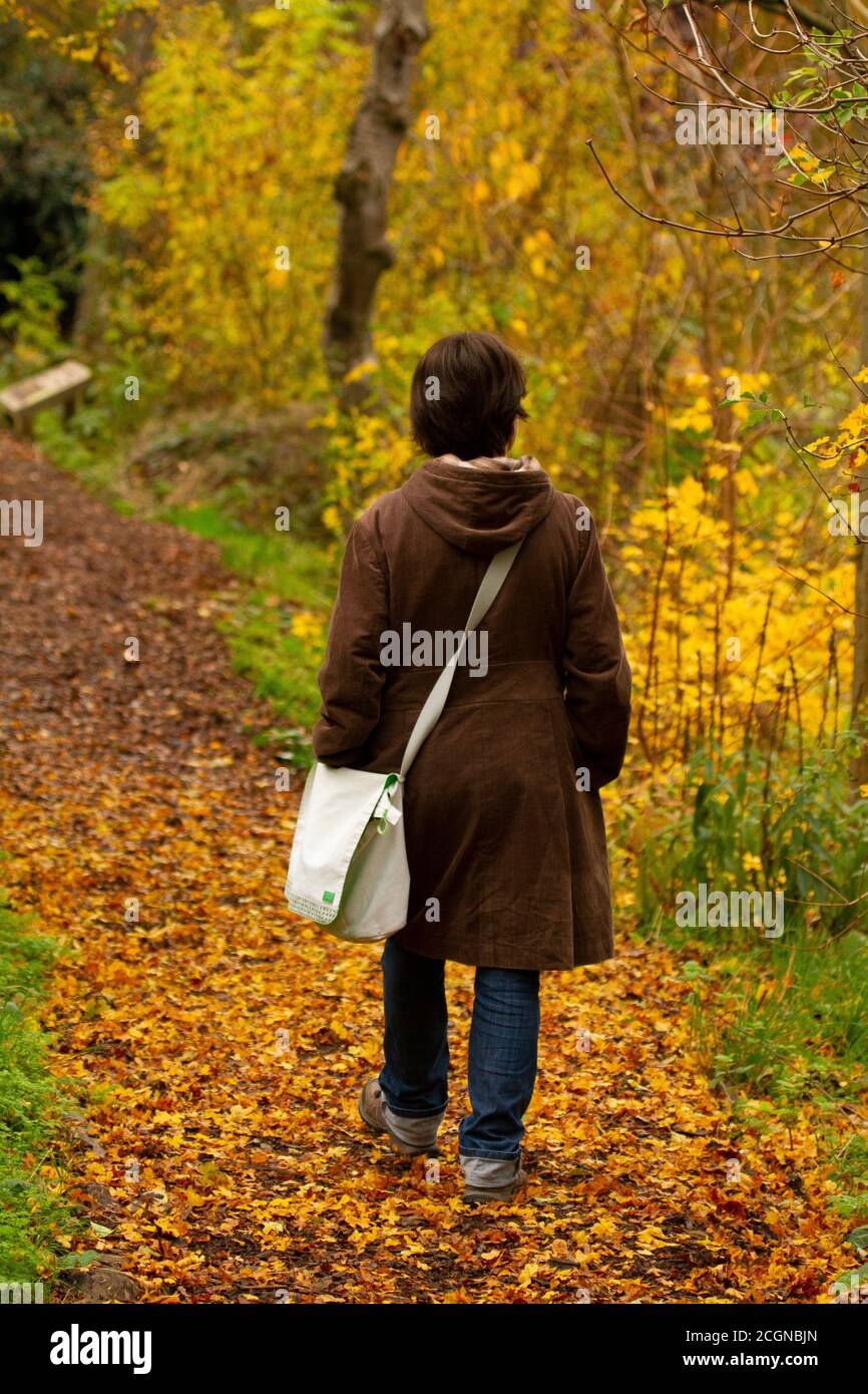 Una giovane donna che indossa un cappotto lungo e una borsa laterale sta camminando da sola su un sentiero nel bosco. Il percorso è coperto da foglie marroni e lei si sta guardando intorno Foto Stock