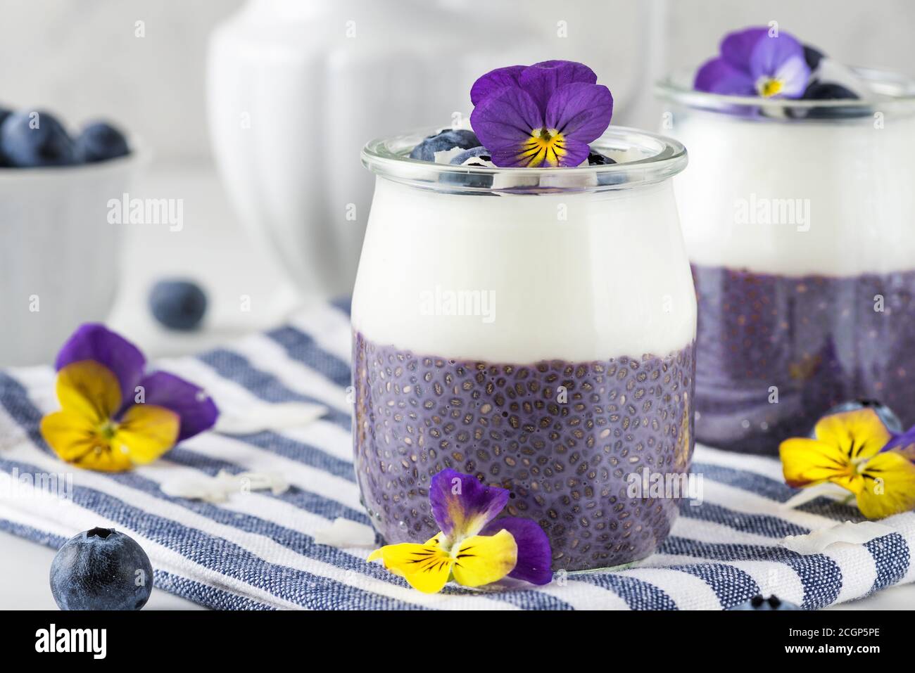 Sana colazione vegana di parfati di mirtillo fatta con frutti di bosco freschi, yogurt al cocco, budino di chia e fiori commestibili in bicchieri su tovagliolo su bianco Foto Stock