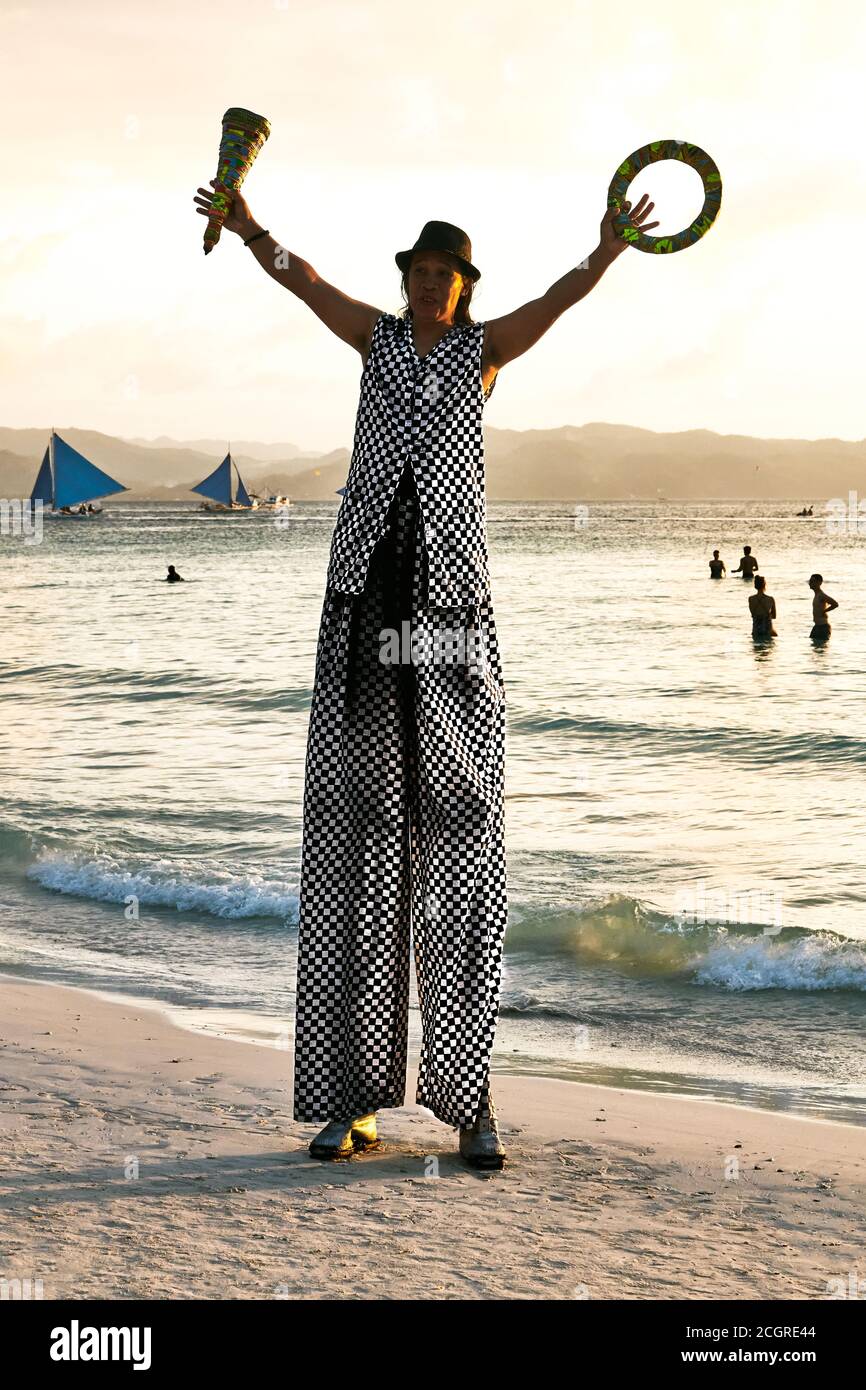 Uomo su palafitte che si giocolano lungo la Spiaggia Bianca al tramonto, con turisti locali come spettatori, poche persone che nuotano e barche a vela blu come sfondo Foto Stock