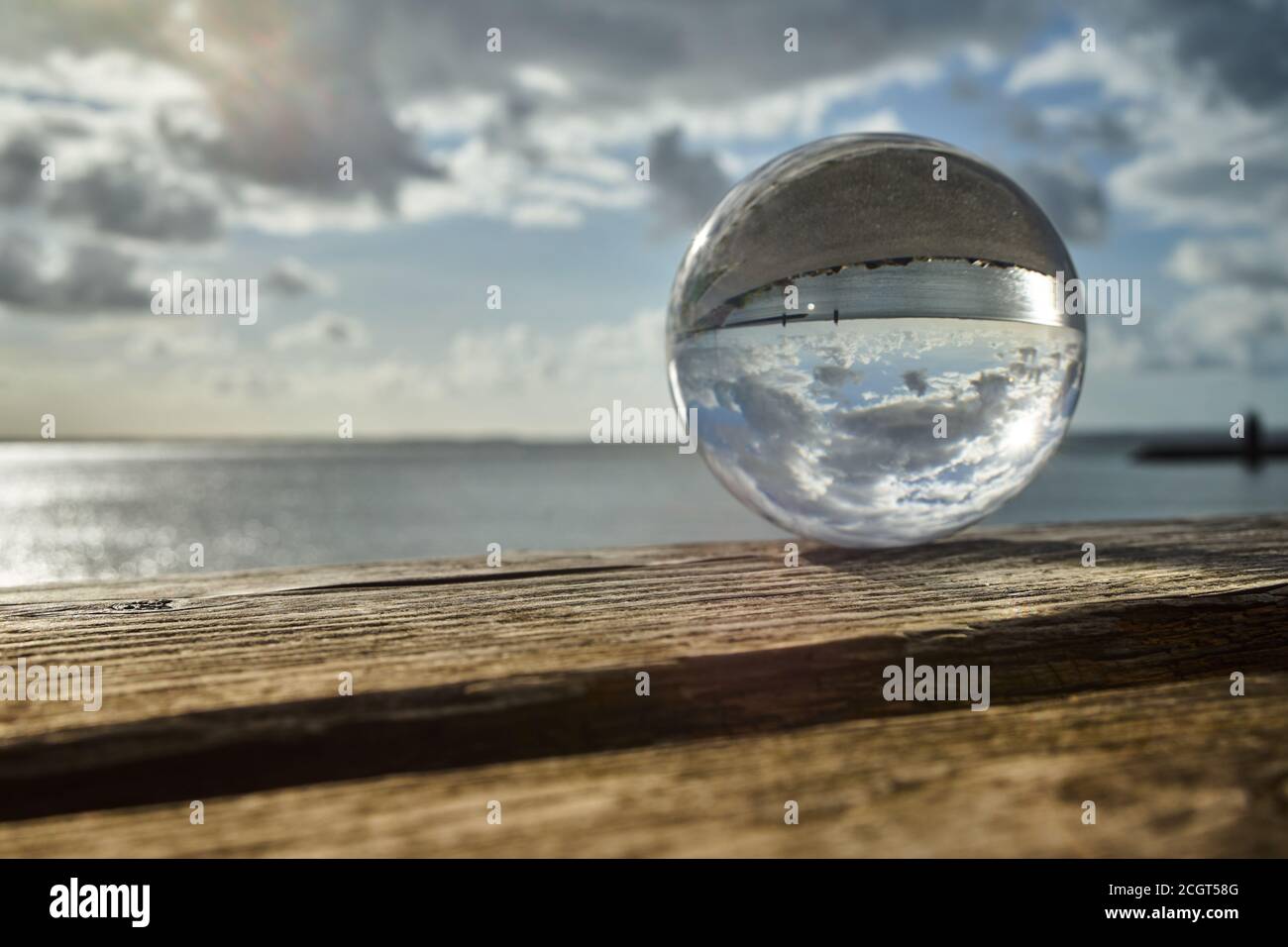 Vista attraverso una sfera di cristallo su un vecchio legno esteso panchina sul mare con le mudflats e il cielo con le nuvole Foto Stock