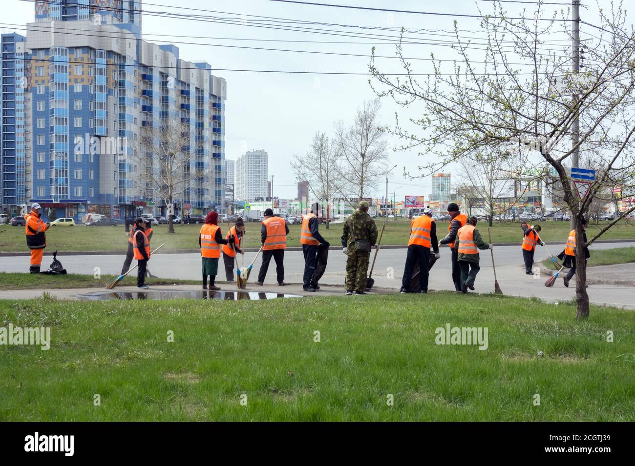 Un gruppo di pulitori di giubbotti protettivi di colore arancione brillante stanno spazzando il marciapiede della città su una strada di primavera in un'area residenziale. Foto Stock