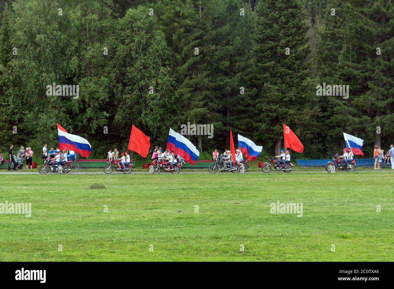 Le moto con bandiere rosse e russe passano davanti agli spettatori sullo sfondo di una foresta di conifere nello stadio. Foto Stock