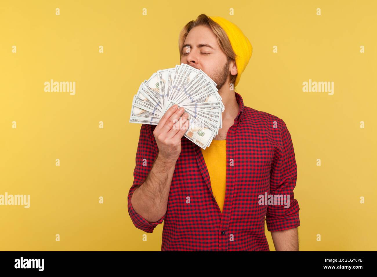 Ricco avaricioso hipster ragazzo in berretto e camicia a scacchi che odora banconote in dollari con espressione di piacere, avido ossessionato con denaro, grande lotto Foto Stock