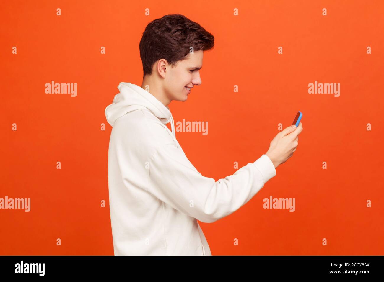 Profilo di felice adolescente sorridente in felpa casual che tiene il telefono cellulare in mani chattando via videochiamata, applicazione di navigazione. Studio al coperto ho girato i Foto Stock