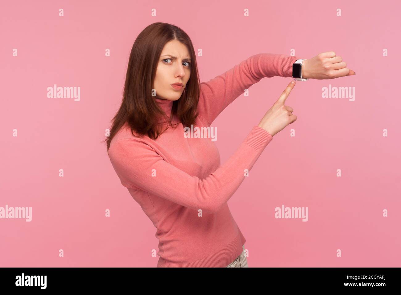 Interessata bruna puntuale donna che punta il dito sullo smartwatch sul polso, guarda il tempo, sbrigati e agisci. Studio al coperto isolato sulla schiena rosa Foto Stock