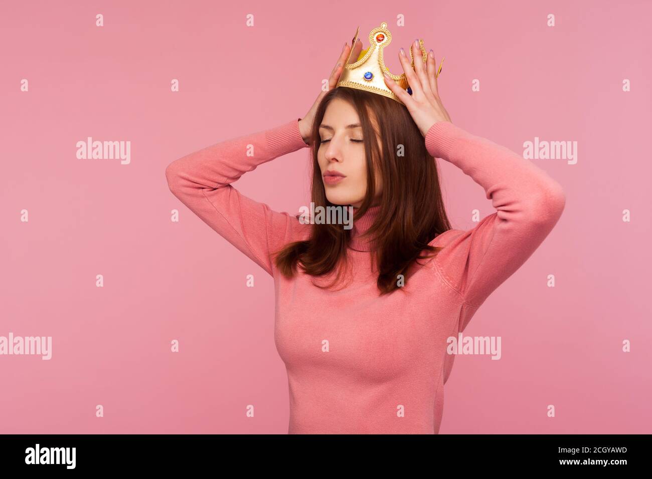 Donna di bruna sicura ed egoista che regola la corona d'oro sulla testa, orgogliosa del suo status, fingendo di essere regina. Studio al coperto isolato su rosa Foto Stock