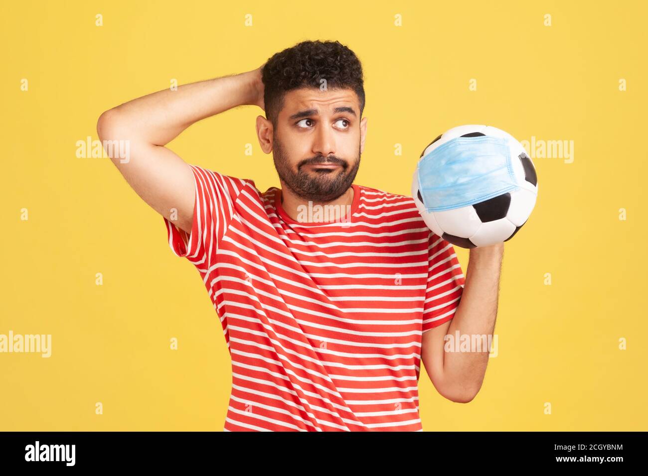 Triste uomo pensieroso in T-shirt rossa che tiene la palla di calcio in maschera protettiva, simbolo della competizione di calcio durante il virus covid-19, punteggio falsificazione. IO Foto Stock