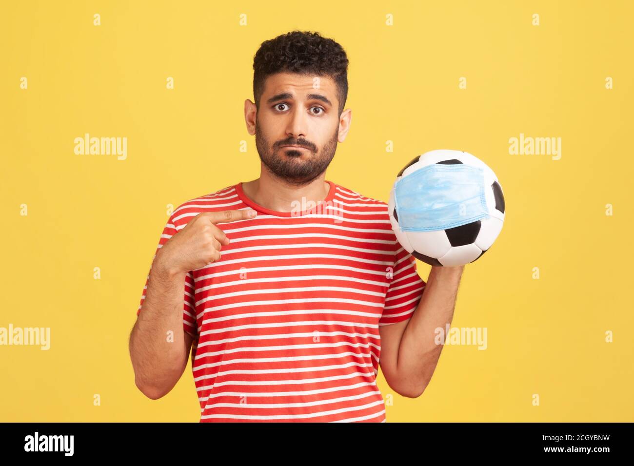 Arrabbiato giovane uomo in T-shirt rossa che punta alla palla in maschera di sicurezza in mano, annullamento di competizioni sportive, isolamento di massa. Studio interno girato isola Foto Stock