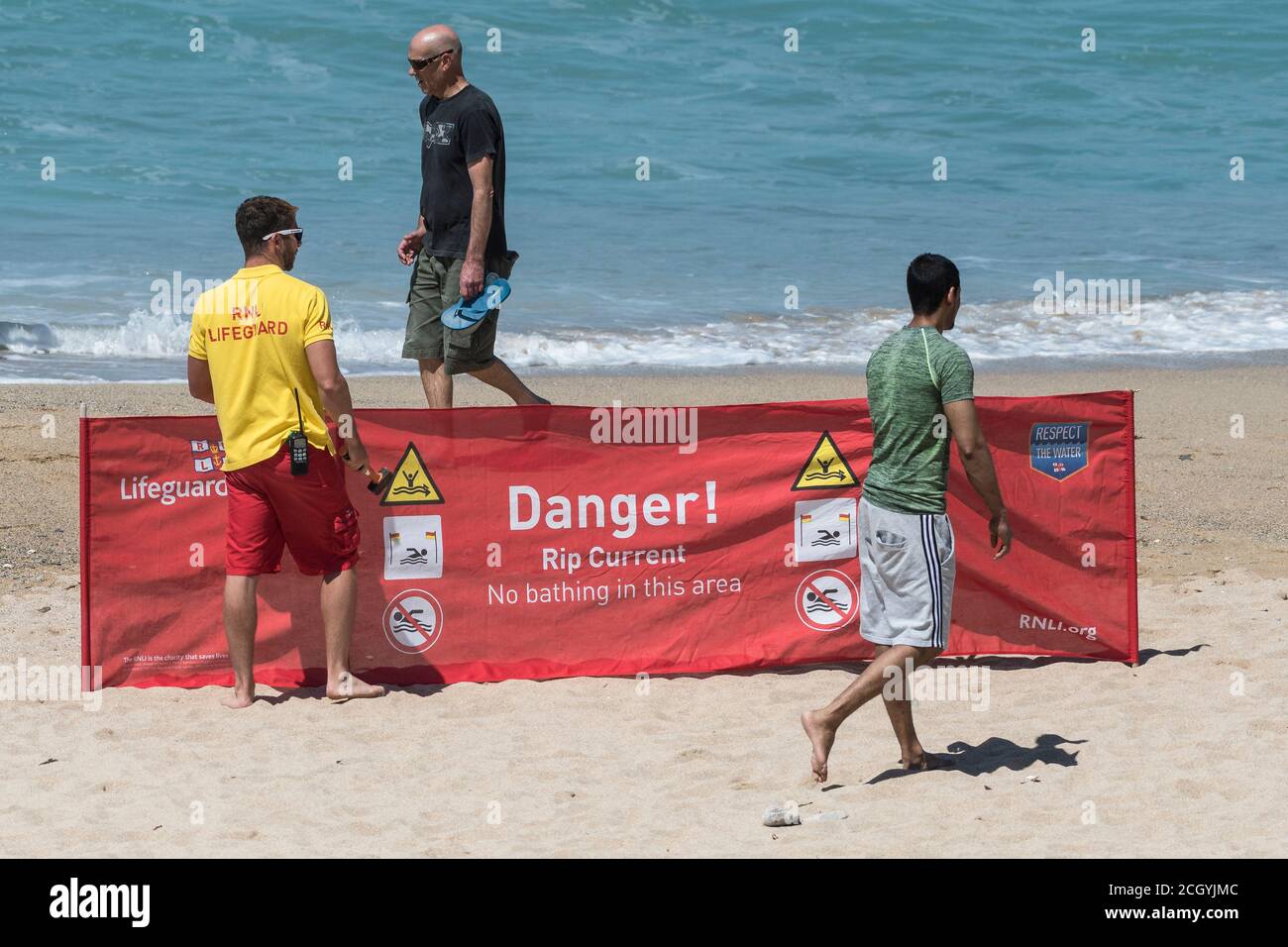 I turisti che camminano davanti a una Lifeguard RNLI, mettono un cartello di avvertimento su Fistral Beach a Newquay in Cornovaglia. Foto Stock