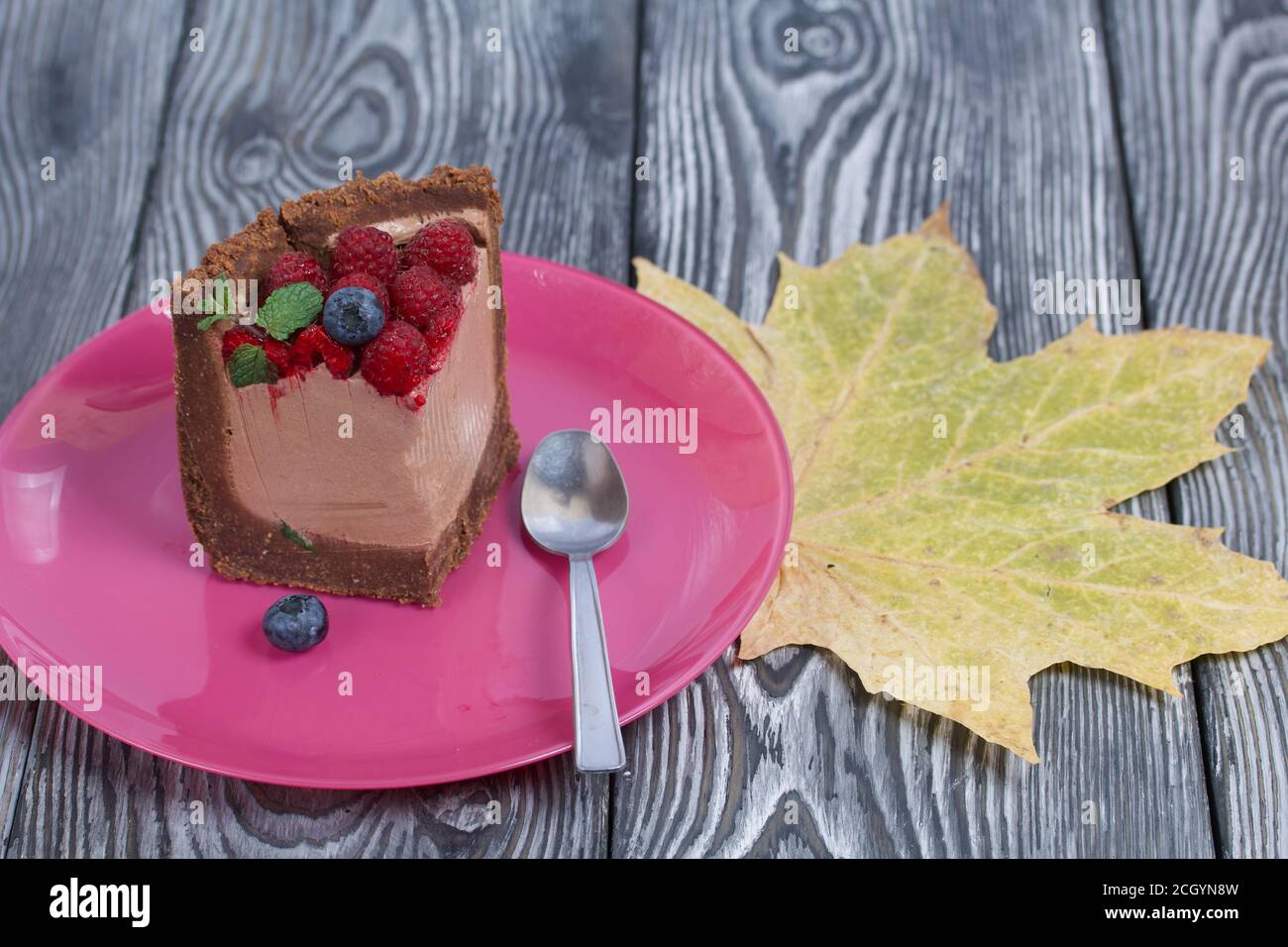 Un pezzo di torta al cioccolato senza forno con formaggio cremoso. Guarnito con lamponi, mirtilli e foglie di menta. Nelle vicinanze si trova una foglia di acero secca. Sul sur Foto Stock