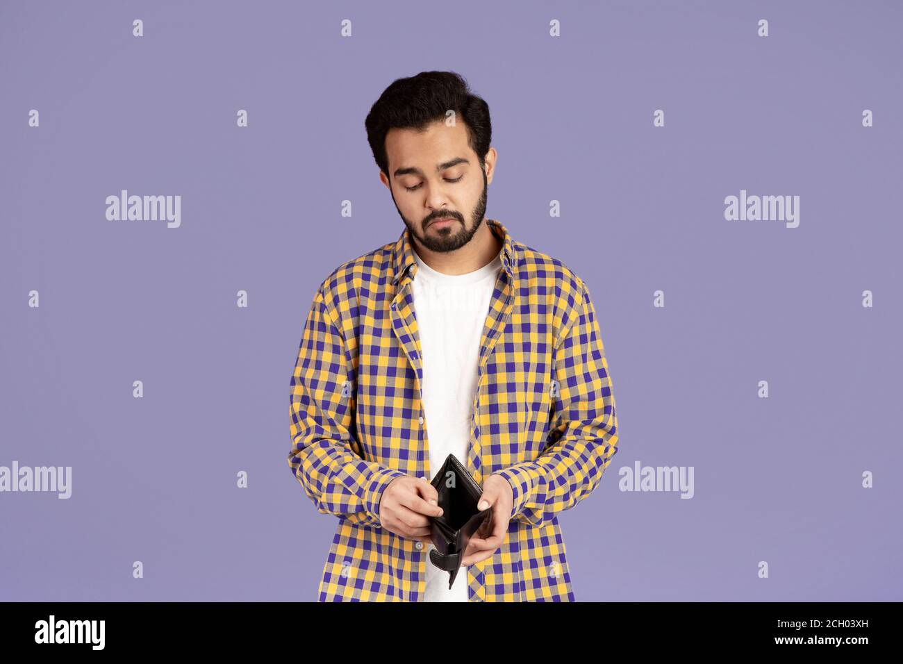 Crisi economica. Insoddisfatto ragazzo indiano che guarda nel portafoglio vuoto su sfondo viola Foto Stock