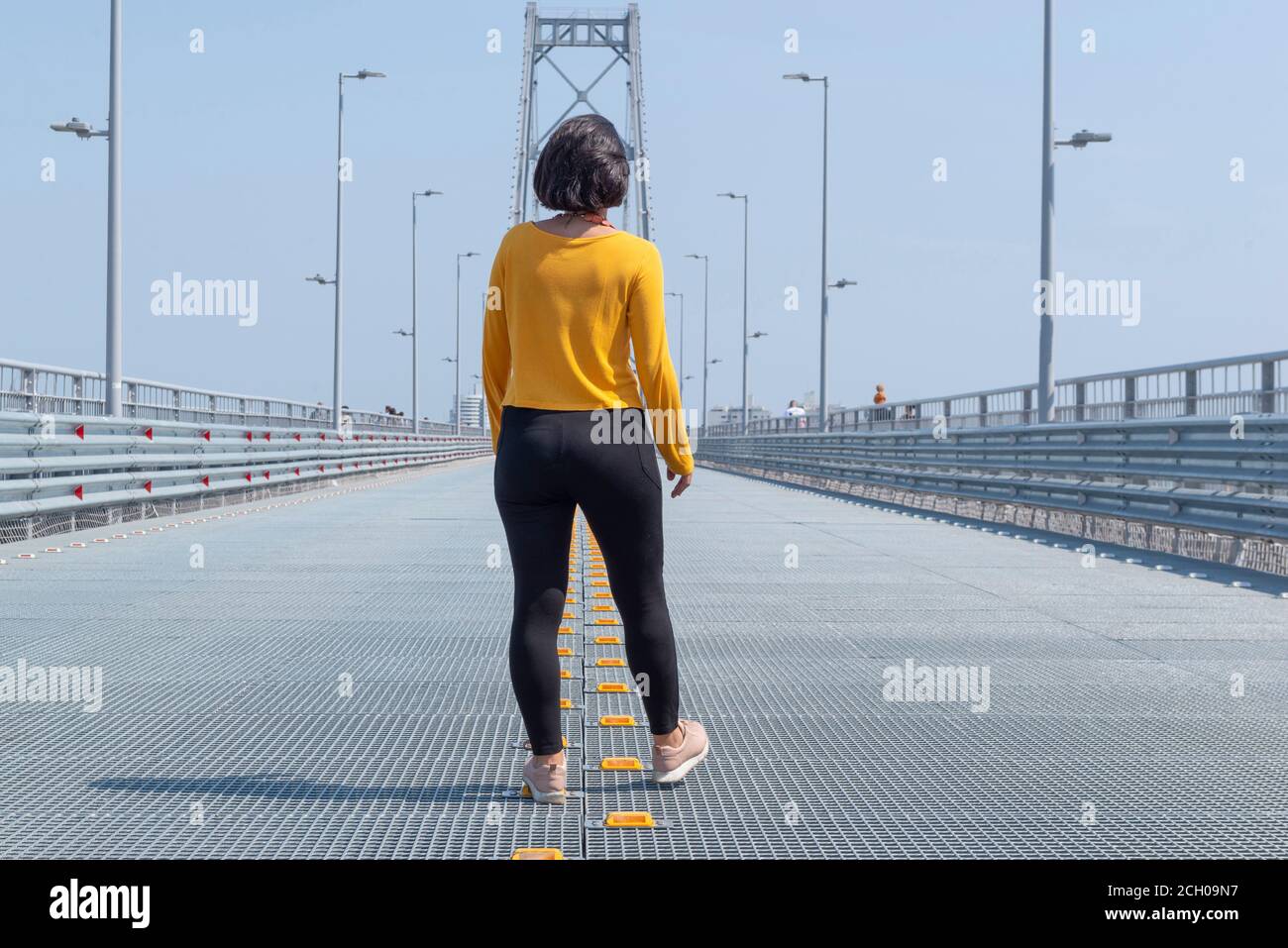 Vista posteriore di donna che cammina nel mezzo di una strada vuota verso il ponte Florianopolis. Ponte in acciaio sotto il bel cielo blu. Continuare a camminare conc Foto Stock