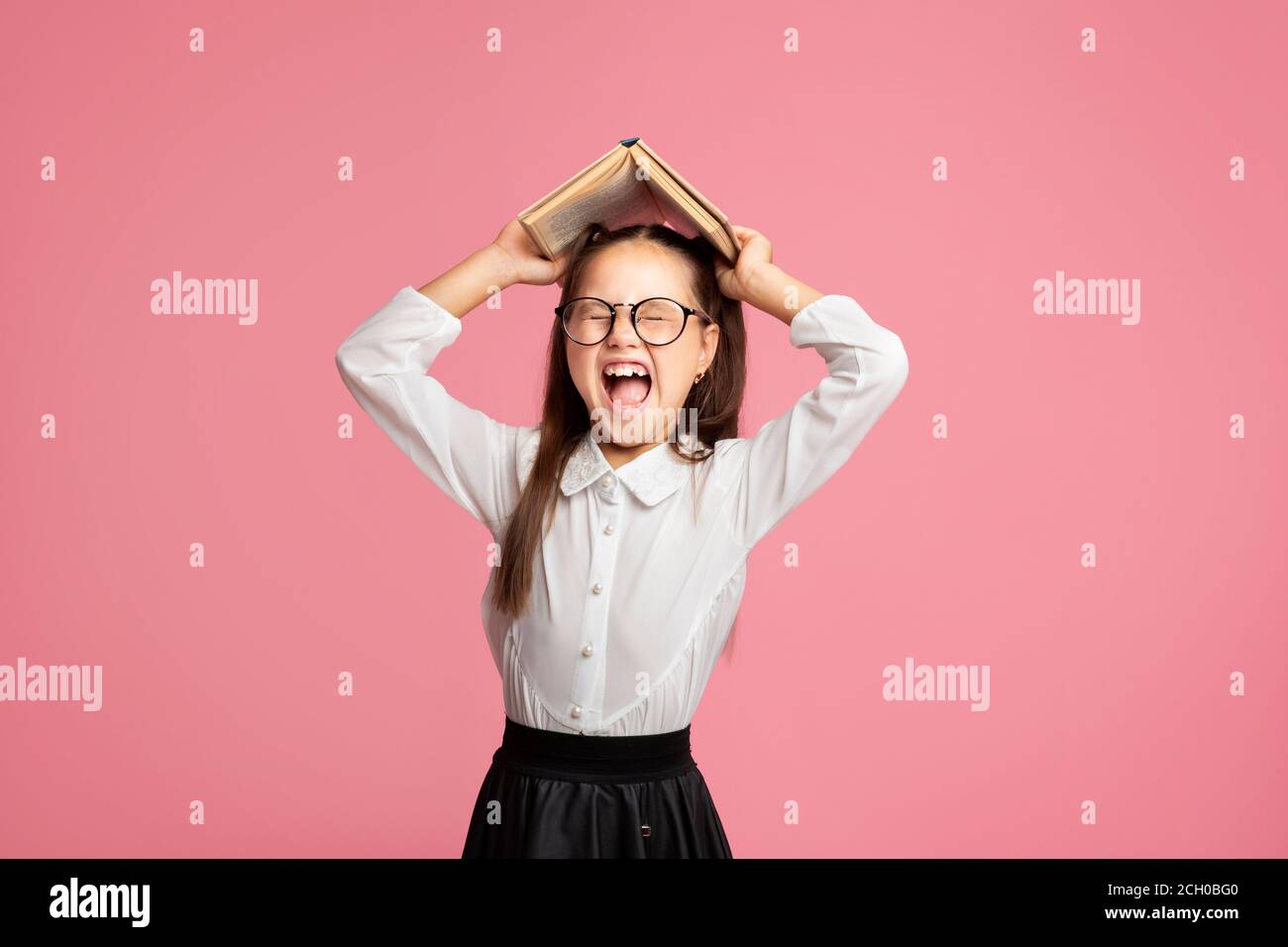 OH no, odio studiare. Studentessa in uniforme e occhiali urla e tiene il libro sulla sua testa Foto Stock