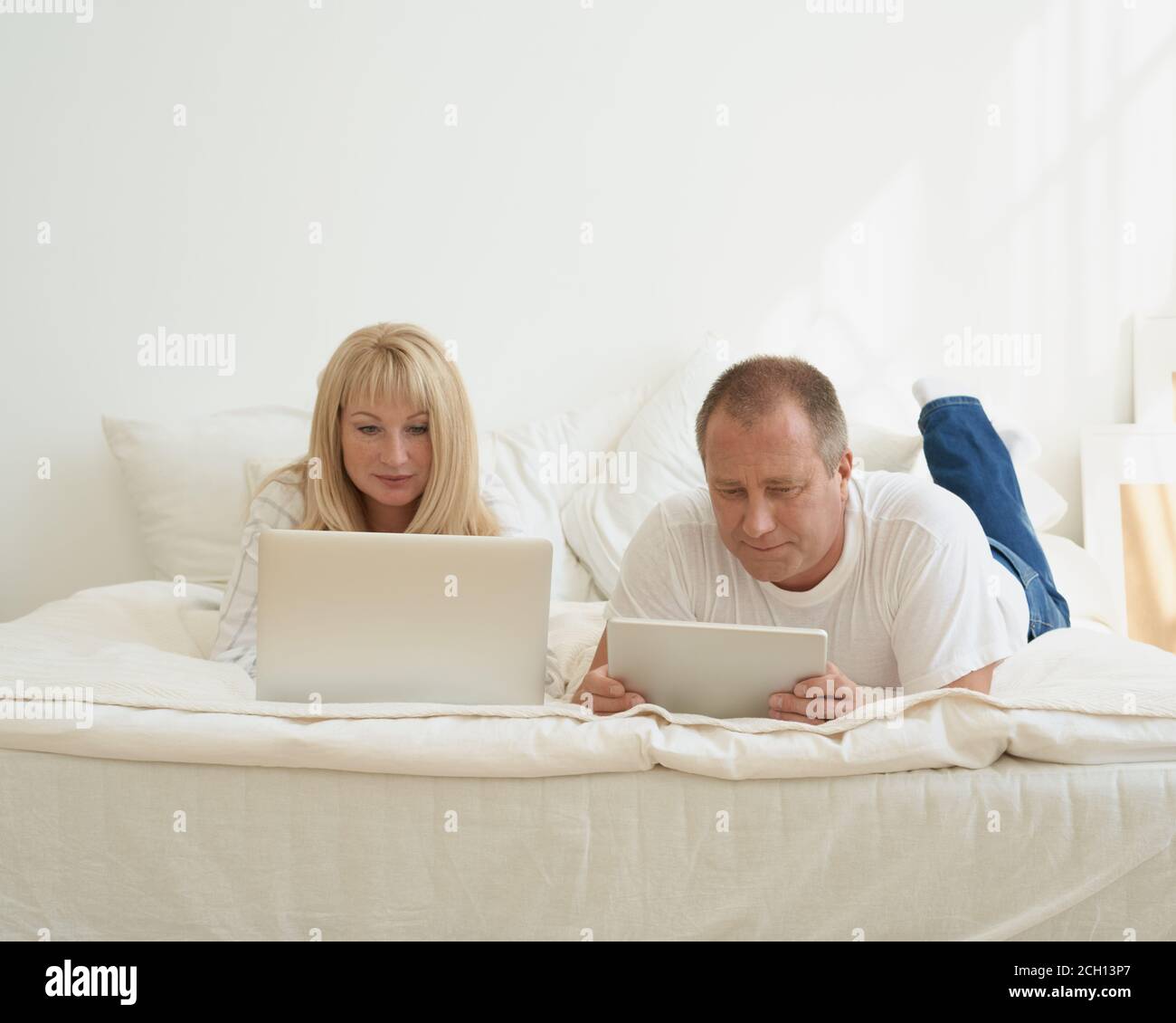 Ritratto di coppia matura con dipendenza digitale a casa. Bell'uomo con un tablet e una bella donna di mezza età con un computer portatile che passa il tempo insieme mentre si sdraiava a letto. Foto Stock
