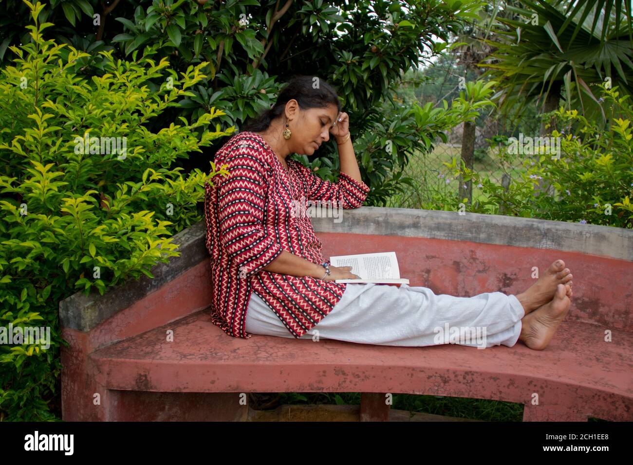 Una bella donna in abbigliamento casual, seduta su una panchina rossa sta leggendo un libro e pensando alla storia in un parco Foto Stock