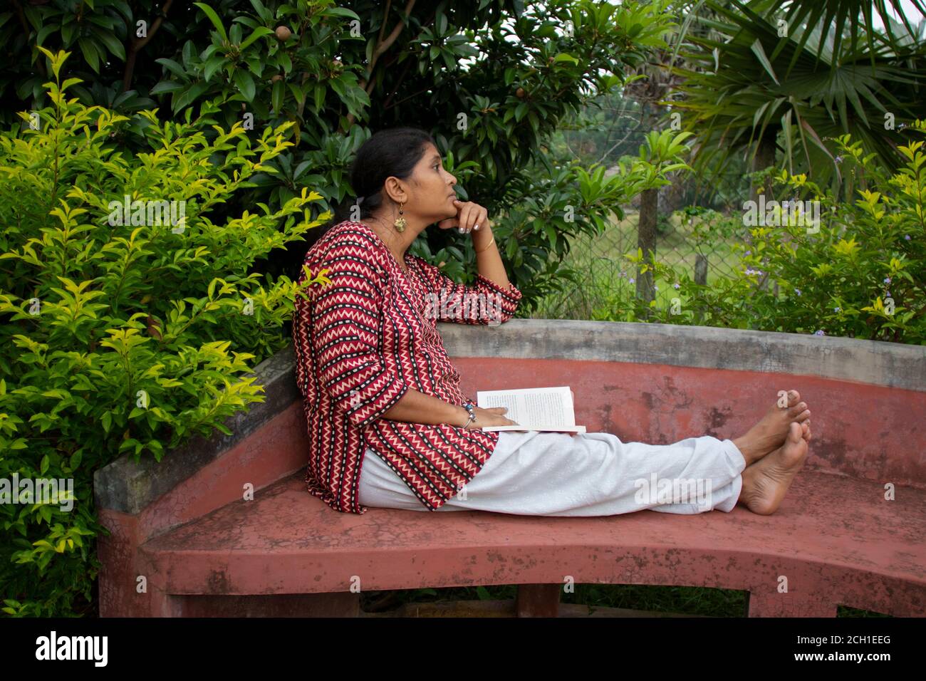 Una bella donna in abbigliamento casual, seduta su una panchina rossa sta leggendo un libro e pensando alla storia in un parco Foto Stock