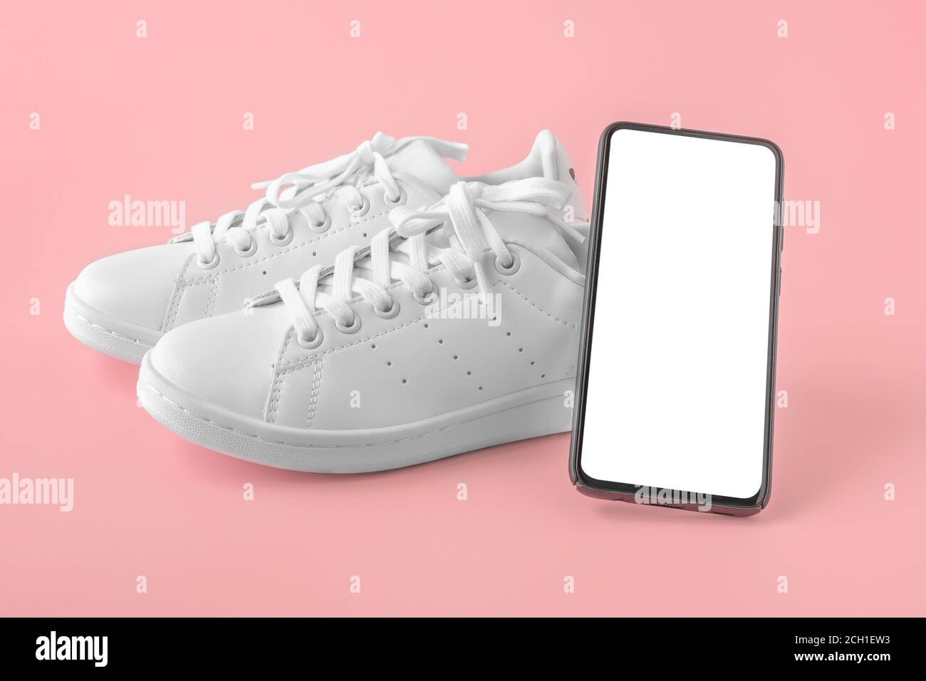 Sneakers e un telefono cellulare con uno schermo bianco vuoto. Telefono cellulare con schermo vuoto, sneakers bianche su sfondo rosa. Derisione. Concetto di app per lo sport Foto Stock