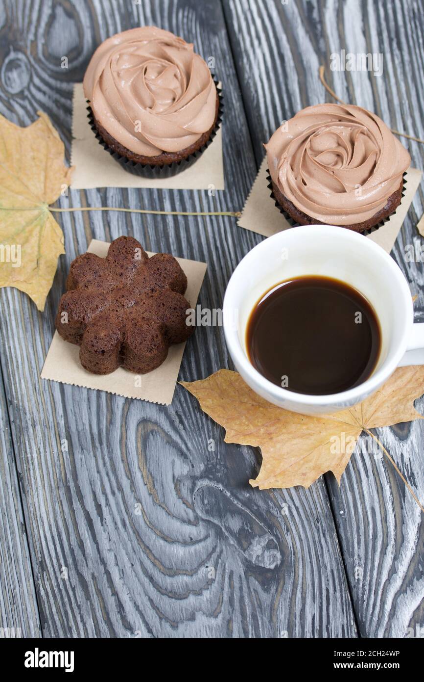 Cupcake al cioccolato con crema di formaggio. Nelle vicinanze si trova una tazza di caffè e foglie di acero essiccate. Su tavole di pino verniciate in bianco e nero. Foto Stock