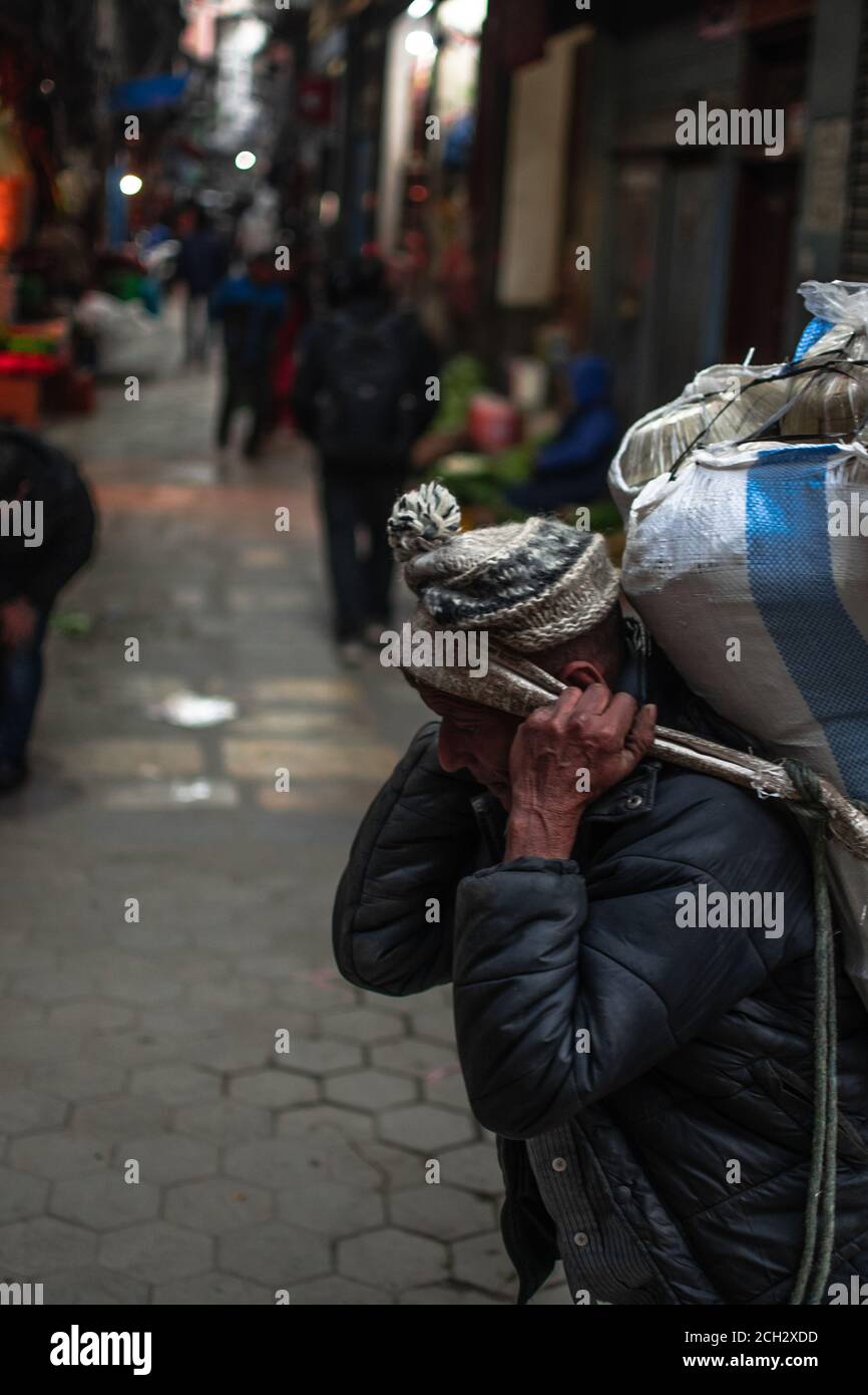 Kathmandu, Nepal - Dicembre 22 2019: Un uomo non identificato con un cappello e una giacca in pelle porta una borsa pesante con la testa a Kathmandu, Dicembre 2019 Foto Stock