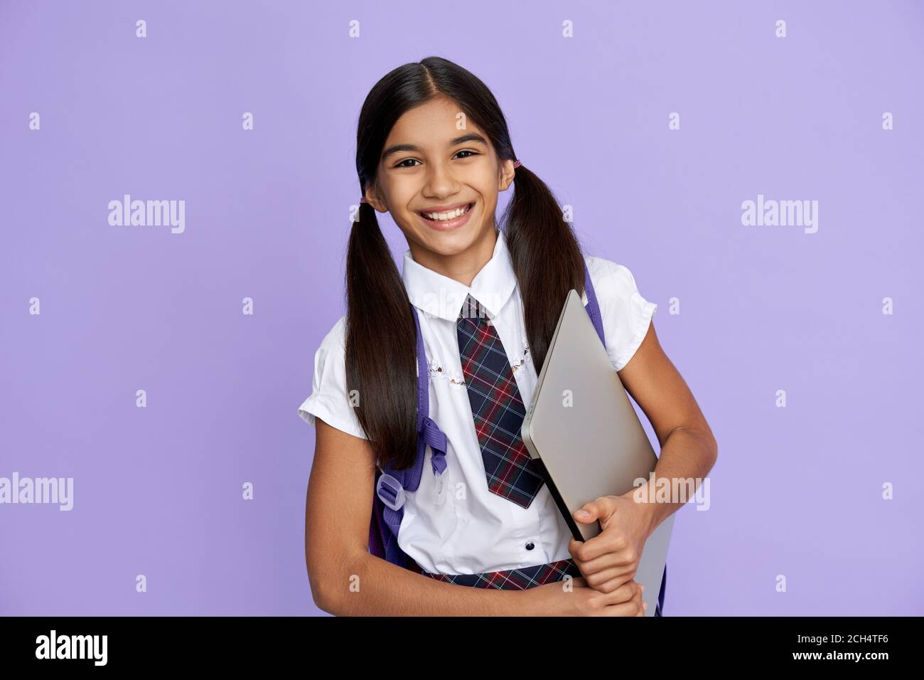 Ragazza indiana felice della scuola che indossa uniforme tenendo il laptop su sfondo lilla. Foto Stock