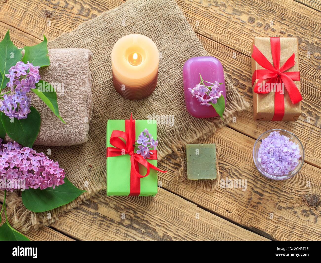 Asciugamani di Terry, sapone e sale marino per le procedure di bagno, candela bruciante, scatole di regalo e fiori di lilla su sackcloth e vecchie tavole di legno. Vista dall'alto. SP Foto Stock