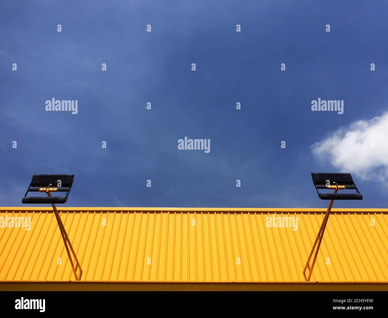 Parete in lamiera di zinco corrugata di colore giallo brillante e tetto con faretto contro il cielo blu. Focus su parete in lamiera di zinco corrugata gialla. Foto Stock