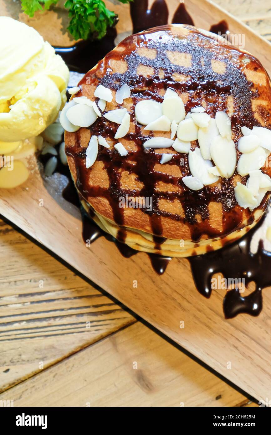 Vista dall'alto dei pancake alla banana con noci di macadamia e sciroppo di cioccolato sulla pila di frittelle e gelato alla vaniglia su un piatto di legno su un tavolo di legno. Foto Stock