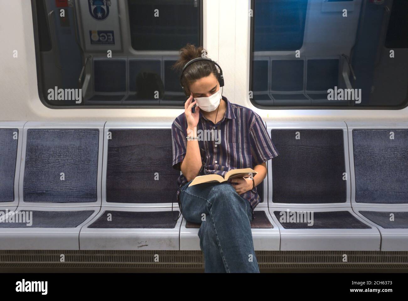 Un giovane adulto isolato e mascherato che ascolta musica e lettura di un libro di letteratura mentre si viaggia in metropolitana Foto Stock