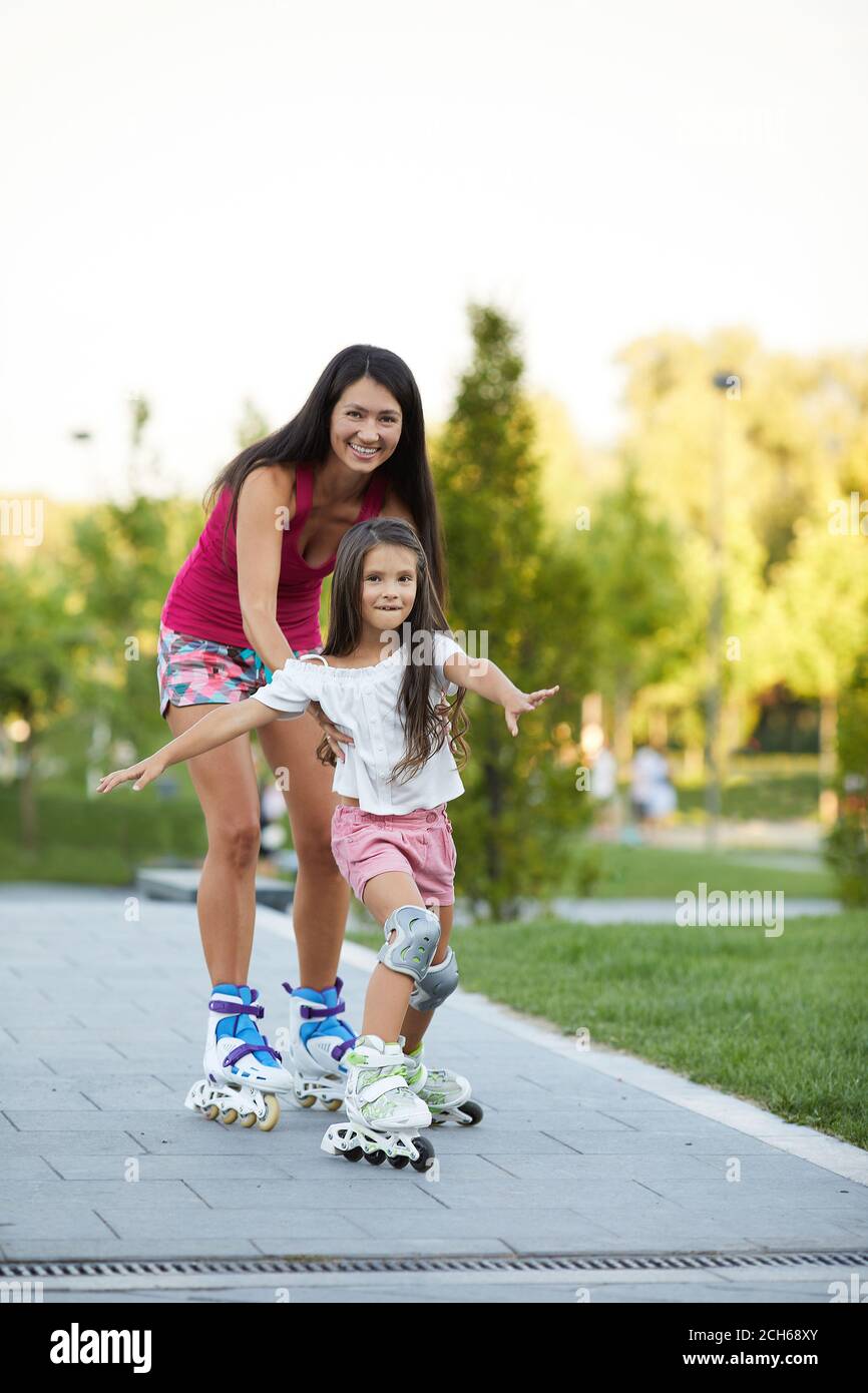 la madre sorridente sta insegnando alla sua piccola figlia a pattinare pattinaggio a rotelle nel parco estivo Foto Stock