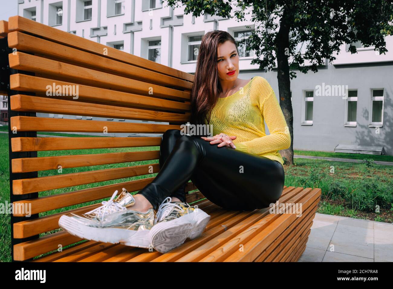 Bella ragazza con lunghi capelli scuri indossa un maglione giallo brillante, lucenti, stretti leggings scuri e sneakers dorate; si siede fuori su una panca di legno Foto Stock