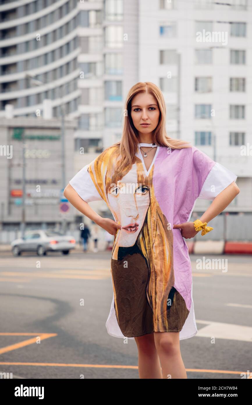 Bella ragazza bionda indossa un vestito colorato con un viso (simile al suo volto) su di esso; sullo sfondo è una strada con auto e edificio alto Foto Stock Bella ragazza bionda indossa un vestito colorato con un viso (simile al suo volto) su di esso; sullo sfondo è una strada con auto e edificio alto Foto Stock