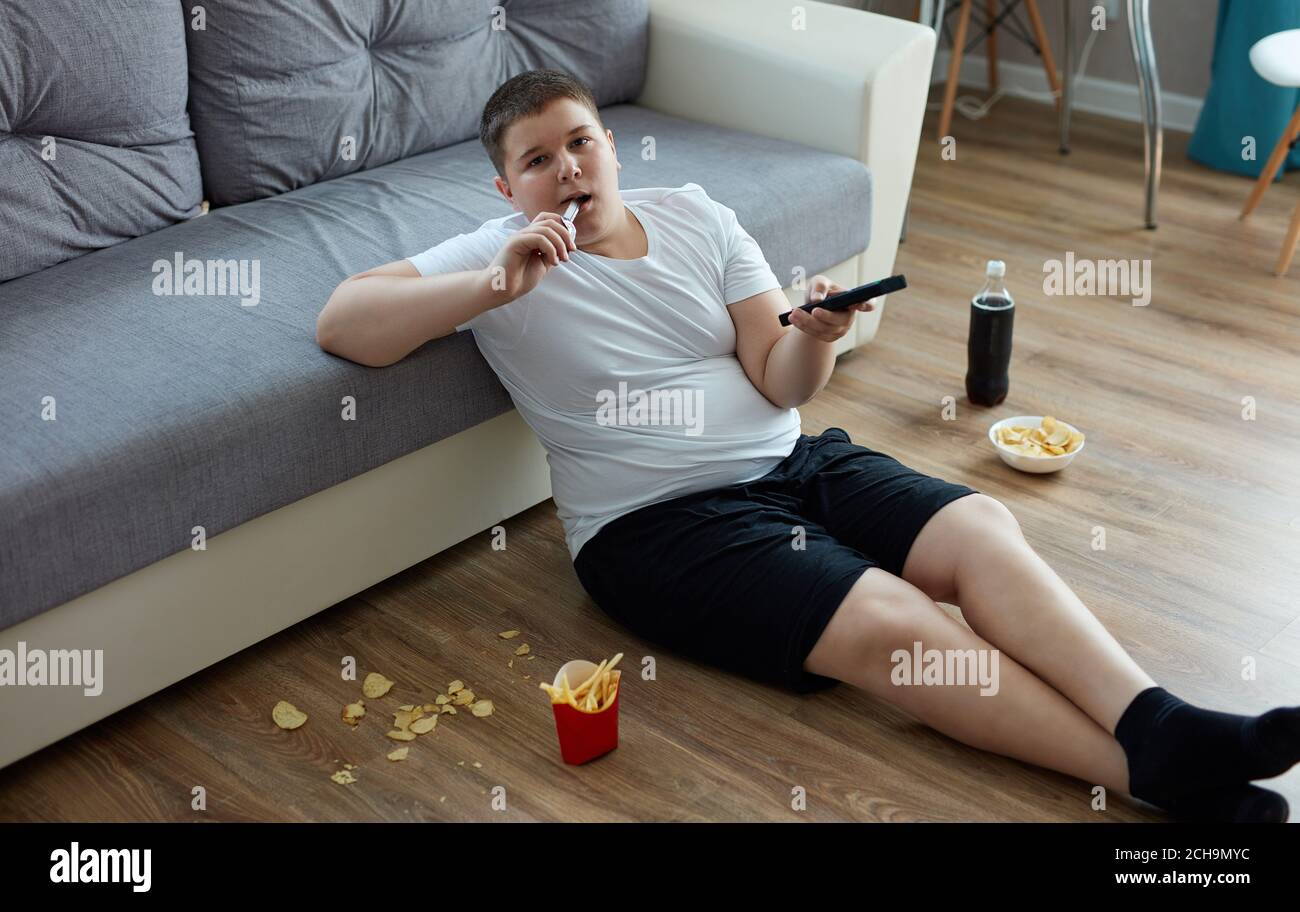 fat boy in sovrappeso mangia cibo spazzatura mentre guardi la tv da solo a casa, siediti sul pavimento con patatine fritte, bevande gassate, patatine. goditi una vita malsana Foto Stock