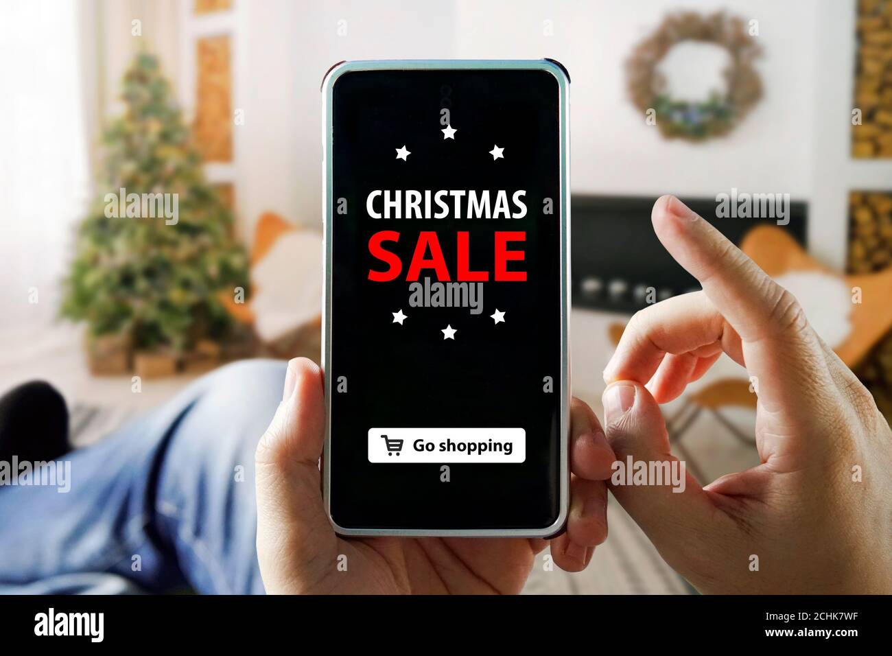 Shopping online di Natale con il telefono. L'uomo si siede in un appartamento e fa acquisti tramite uno smartphone in un negozio online. Albero di Natale, regali Foto Stock