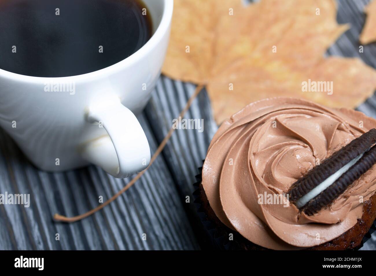 Cupcake al cioccolato con crema di formaggio. Nelle vicinanze si trova una tazza di caffè e foglie di acero essiccate. Su tavole di pino verniciate in bianco e nero. Foto Stock