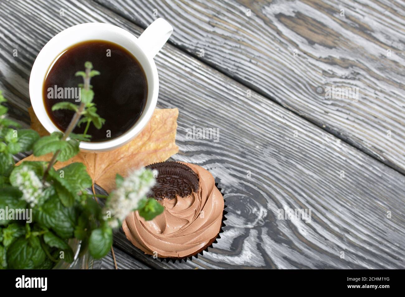 Cupcake al cioccolato con crema di formaggio. Nelle vicinanze si trova una tazza di caffè e foglie di acero essiccate. C'è un vaso con ciuffi fioriti di menta. Foto Stock