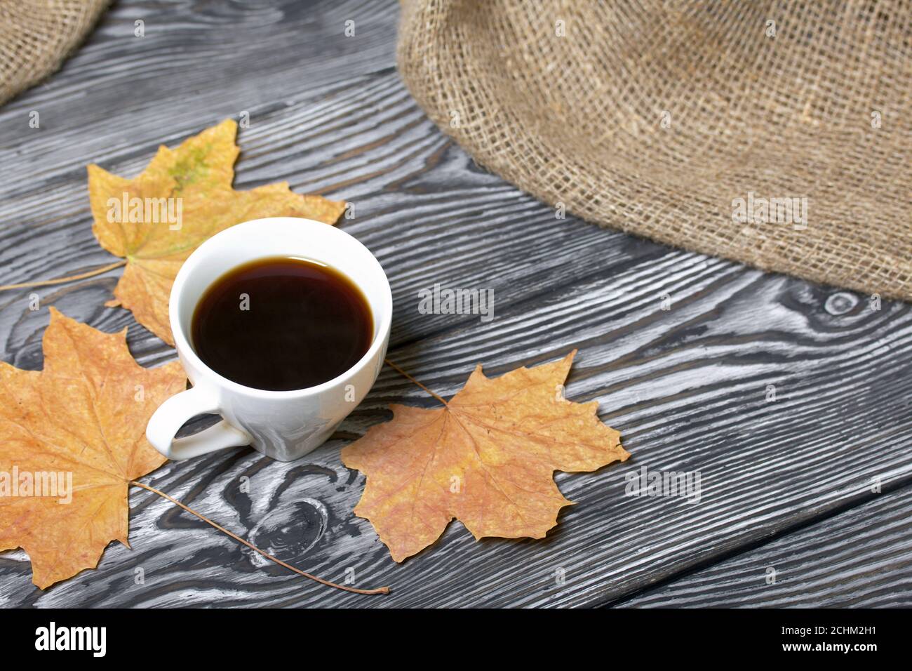 Una tazza di caffè tra foglie di acero secche. Su tavole di pino verniciate in bianco e nero. Nelle vicinanze si trova un panno di lino ruvido. Foto Stock