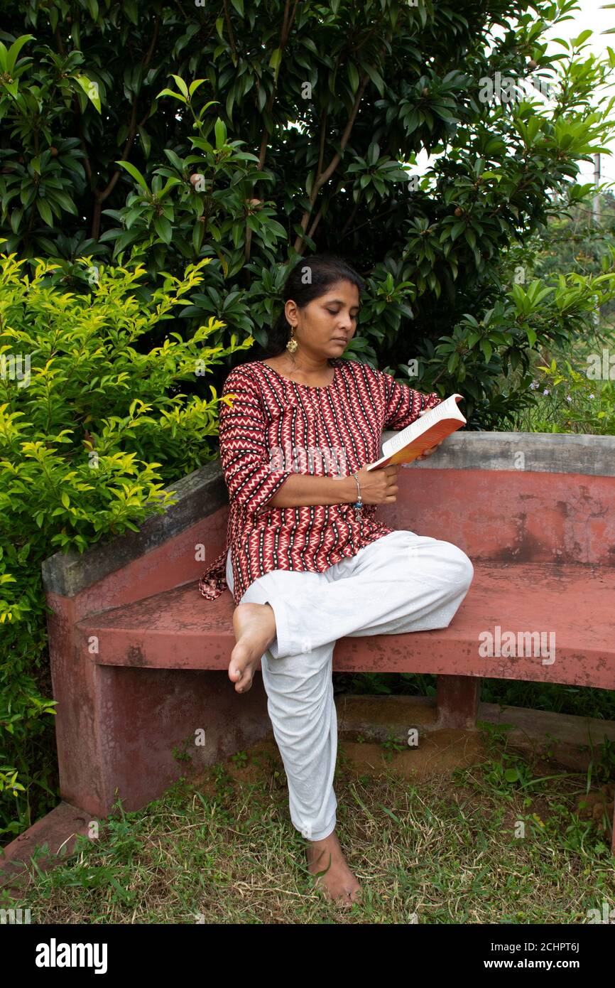 Una bella donna in abbigliamento casual, seduta su una panchina rossa sta leggendo un libro e pensando alla storia in un parco Foto Stock