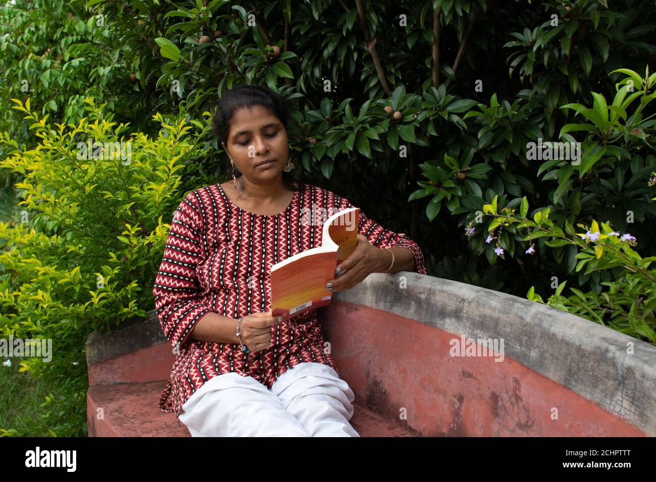 Una bella donna in abbigliamento casual, seduta su una panchina rossa sta leggendo un libro e pensando alla storia in un parco Foto Stock